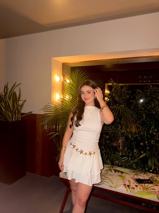 A woman in a white sleeveless star-embroidered dress poses indoors near potted plants and warm wall lighting.