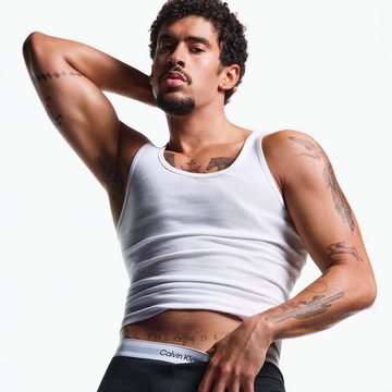 A young man with curly dark hair, a goatee, and visible tattoos on his arms poses confidently in a white Calvin Klein tank top, with the "Calvin Klein" text visible on the waistband of his black underwear. The image accompanies an article suggesting he is an early favorite for PEOPLE's Sexiest Man Alive based on Polymarket predictions.