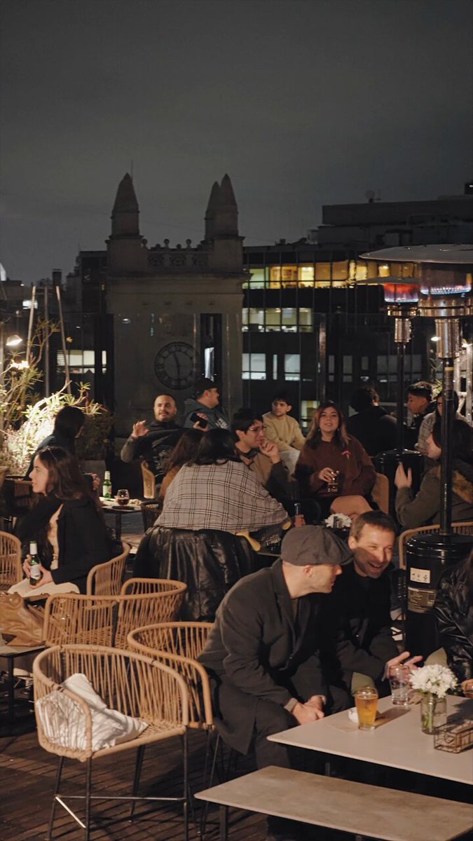 The image shows a group of people gathered around tables, enjoying each other's company at an outdoor restaurant or bar. The atmosphere appears lively and social as they engage with one another over drinks and food. There are several chairs and dining tables scattered throughout the scene, indicating that this is likely a popular spot for patrons to relax and dine al fresco.

The photo also includes some interesting elements such as a clock on the wall in the background, which adds an urban touch to the setting. Additionally, there's a handbag placed near one of the tables, suggesting that someone has recently arrived or is preparing to leave. The presence of multiple people and various dining arrangements creates a sense of community and shared enjoyment among the patrons at this outdoor venue.