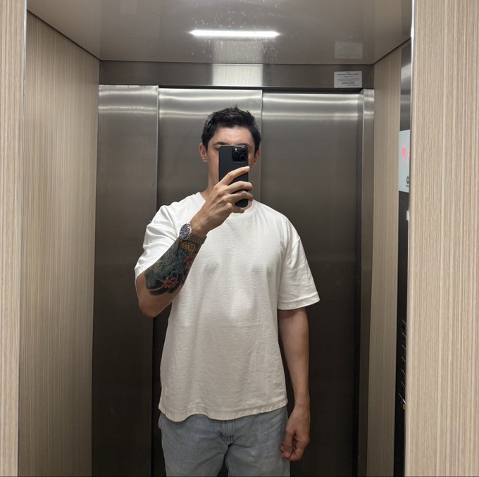 A mirror selfie of a man with short dark hair wearing a white t-shirt and grey pants, showing a tattooed arm, standing in an elevator.