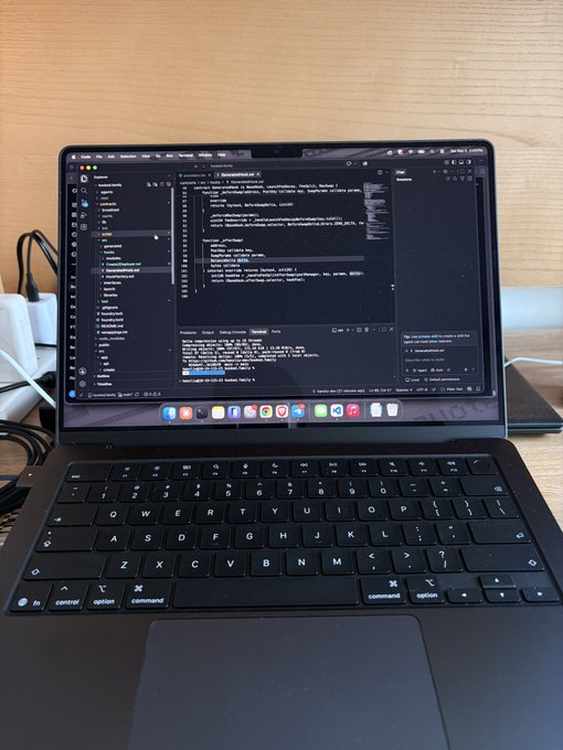 A laptop displaying a code editor (appears to be VS Code) with various files open and a dark theme, sitting on a wooden desk with cables visible.