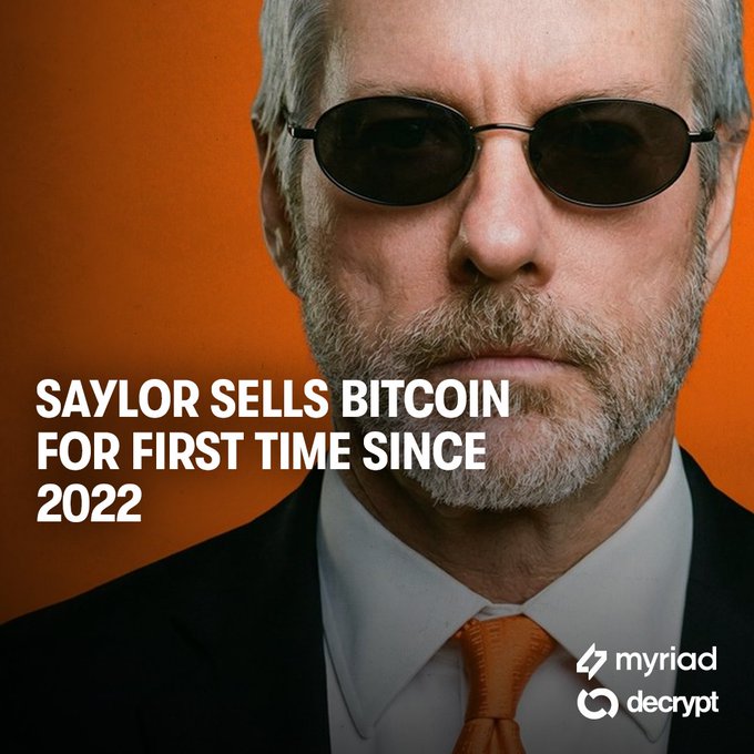 The image features a man with a white beard wearing a black suit and sunglasses and holding a camera. The caption reads "MORNING UPDATE: Bitcoin bleeds after Saylor sells, falls another 3.7% to $69,300" and provides information about some market updates in the cryptocurrency world.