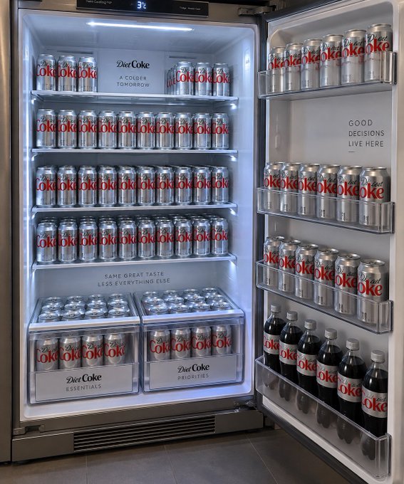 An open refrigerator stocked with Diet Coke cans and bottles, with branding text including 'Diet Coke A COLDER TOMORROW', 'GOOD DECISIONS LIVE HERE', 'SAME GREAT TASTE LESS EVERYTHING ELSE', 'Diet Coke ESSENTIALS', and 'Diet Coke PRIORITIES'.