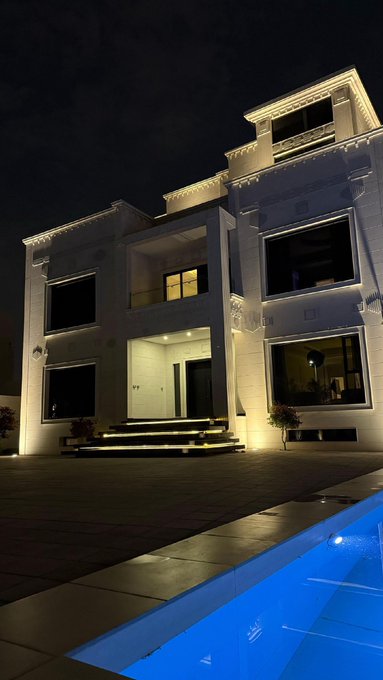Nighttime photograph of a modern white multi-story villa with illuminated architectural details and large windows, featuring a swimming pool with blue-lit water in the foreground.
