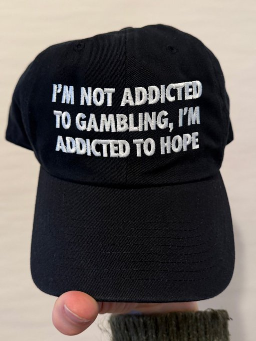 A black baseball cap held up by a hand, with embroidered white text reading "I'M NOT ADDICTED TO GAMBLING, I'M ADDICTED TO HOPE."