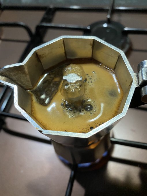 A top-down view shows espresso brewing in a silver octagonal Moka pot on a gas stovetop, with rich crema forming as coffee bubbles up through the spout.