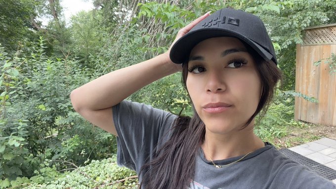 A young woman wearing a dark cap with "OKX" branding and a gray t-shirt poses outdoors with her hand resting on her hat, surrounded by lush greenery. The OKX-logoed cap directly ties into the post's reference to the cryptocurrency exchange, showcasing brand affiliation in a casual weekend selfie.