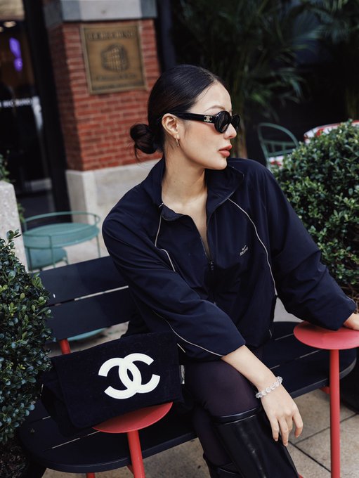 Stylish photograph of a woman wearing a navy blue jacket, dark sunglasses, and black knee-high boots, seated on a red and black bench outdoors holding a black Chanel handbag.