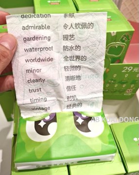 A hand holding a folded paper napkin printed with English words and their Chinese translations (such as 'dedication', 'admirable', 'gardening'), positioned in front of green blind box packaging priced at '29元'.