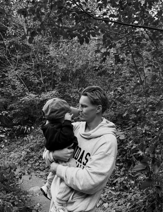 A black-and-white photo of a man in a "ADIDAS" hoodie holding a small child wearing a cap in a wooded area.