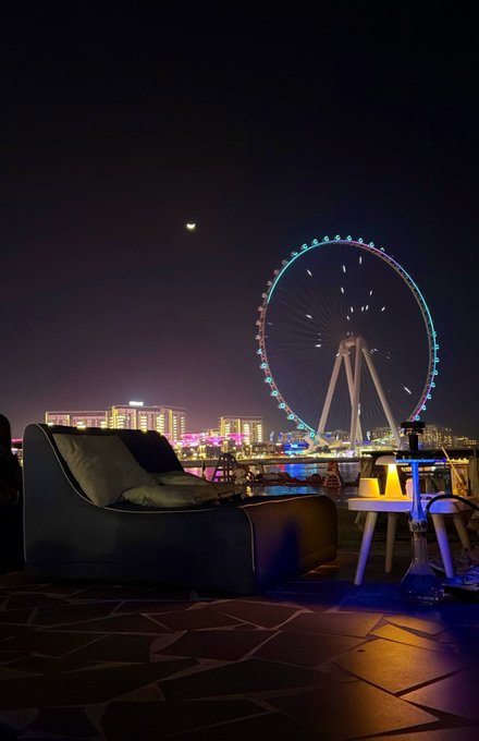 A nighttime view from a rooftop lounge features a couch and small table in the foreground, with an illuminated Ferris wheel and city skyline in the background under a crescent moon. The "see you at the flip" caption suggests a transition tied to the rotating wheel and the turning of day to night, symbolizing a new dawn for the crypto community.