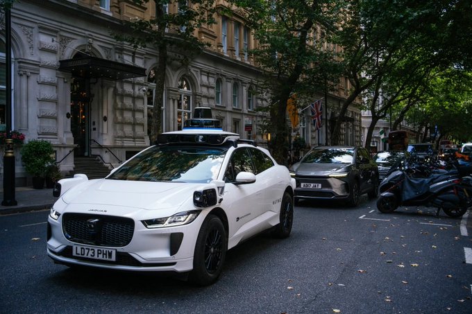 A white Waymo-branded Jaguar self-driving car with roof sensors drives on a city street, with a UK license plate reading 'LD73 PHW.'