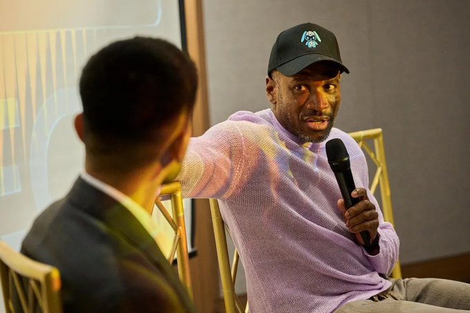 A man wearing a black baseball cap with a small logo and a light purple sweater is speaking into a microphone during what appears to be an interview or panel discussion.