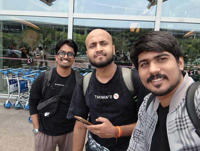 Three men pose for a selfie outside an airport terminal, with the central figure wearing a black shirt displaying the text 'TSIPRASIAST' (which appears mirrored).