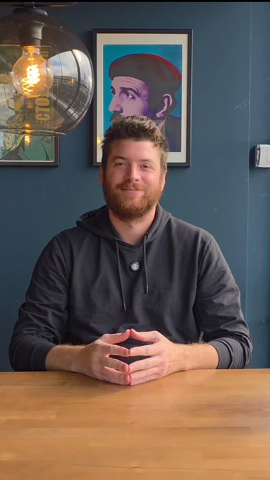 MrBeast sits at a wooden table with his hands folded, smiling slightly at the camera in front of a dark blue wall decorated with framed portraits and a hanging light bulb.