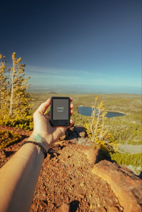 A hand holds a CORZIO hardware wallet displaying a "start setup" screen while overlooking a scenic mountain landscape with a lake from a rocky cliff. The image visually reinforces the post's message about crypto OPSEC by placing the hardware wallet in an isolated, outdoor setting—suggesting that secure crypto practices begin with careful device selection and controlled environments before any transaction is signed.