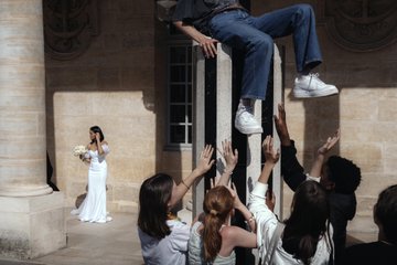 A group of people reach up to support a man in jeans and white sneakers sitting on a tall pillar, while a bride in a white dress watches from the left.