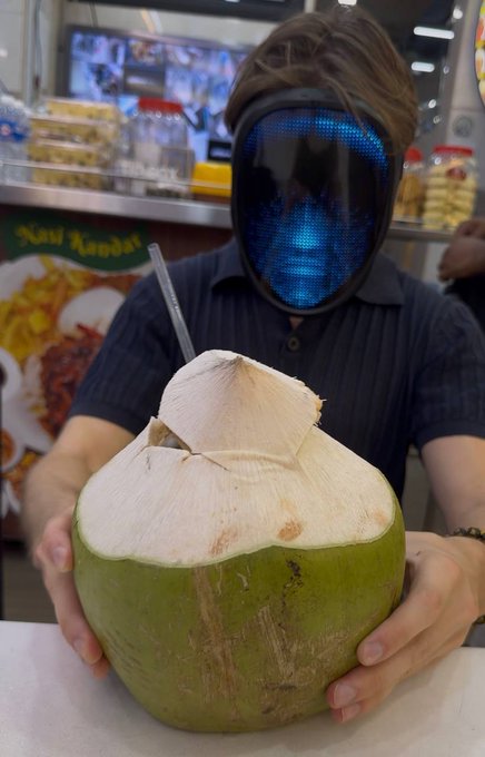 A person wearing a digital face mask holds up a fresh green coconut with a straw at what appears to be a Singapore hawker stall, with "Nasi Kandar" signage visible in the background — a perfect "GM Singapore" greeting showcasing a classic local refreshment.