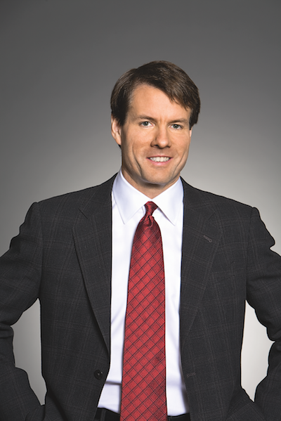 Professional headshot of a man in a dark checkered suit, white shirt, and red patterned tie, smiling against a neutral gray background.