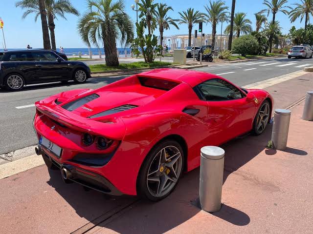 A red Ferrari sports car parked on a palm-lined street with the Mediterranean Sea in the background.