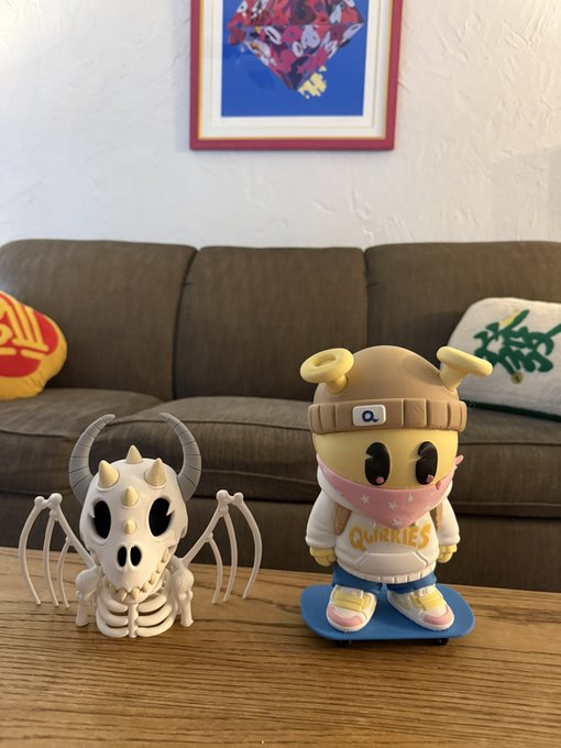 Two designer toy figures—a horned skeletal creature and a character labeled 'QUIRKIES' wearing a beanie and bandana—sit on a wooden coffee table in front of a brown couch.