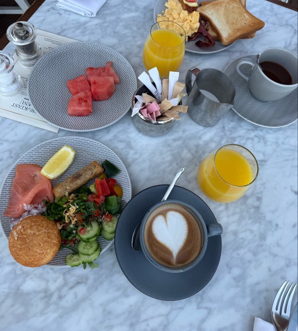 An overhead view of a breakfast spread on a marble table, featuring a plate of fresh fruit including watermelon, a savory plate with salmon and vegetables, coffee with heart latte art, orange juice, toast, and assorted pastries.