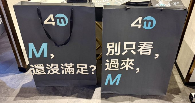 Two large dark gray gift bags from 4M exchange featuring bilingual slogans—"M, 還沒滿足?" (M, still not satisfied?) and "別只看, 過來, M" (Don't just look, come over, M)—promote the crypto exchange's lively, casual event at Token2049.