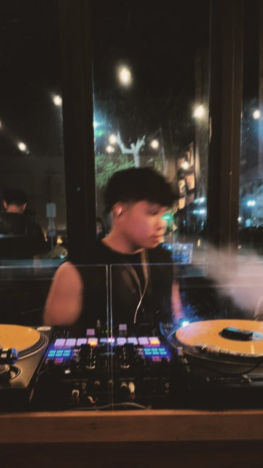 A blurry photo of a young boy with short dark hair working at a DJ mixing board with turntables, visible through a window at night.