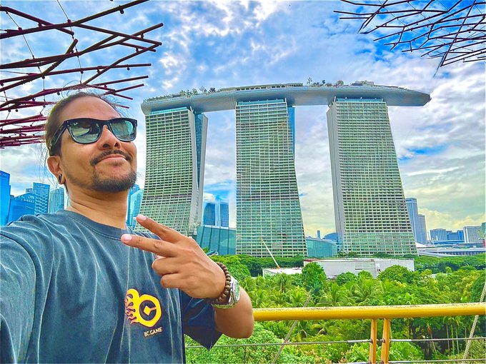 A man with sunglasses poses in front of the Marina Bay Sands hotel in Singapore, making a peace sign while wearing a blue t-shirt.