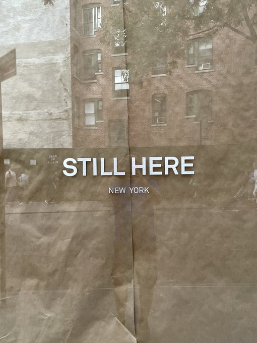 A crumpled newspaper poster taped to a street pole displays the text "STILL HERE" and "NEW YORK" over a faded urban street scene, with a brick building visible behind the translucent paper. The post highlights discovering this mysterious NFT-related artwork in the real world, transforming digital art into a tangible urban treasure hunt.