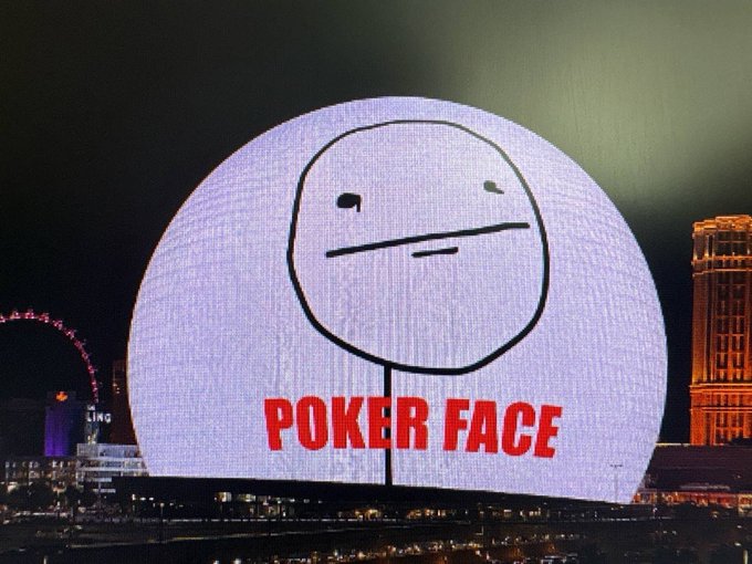 A large spherical LED display at night shows a simple line-drawn neutral face above the red text 'POKER FACE', with a Ferris wheel and illuminated buildings visible in the background cityscape.