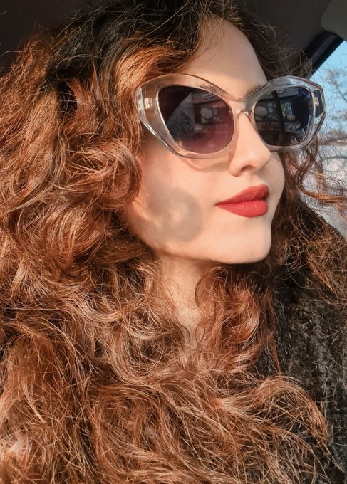 A close-up selfie of a woman with long auburn curly hair wearing cat-eye sunglasses and bold red lipstick, taken from inside a vehicle.
