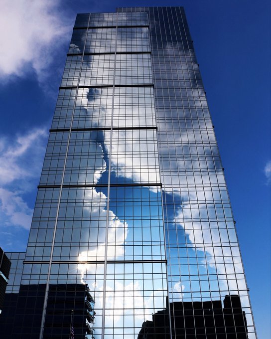 A low-angle architectural photograph of a tall modern skyscraper with a dark blue glass facade reflecting the bright sky and white clouds.