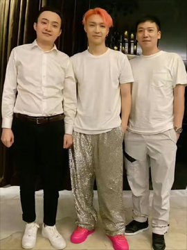 I cannot identify the specific individuals in this photo. I can describe that it shows three men posing together — one in a white button-up shirt, one in the center wearing a white t-shirt and sparkly silver pants with pink hair, and one in a cream-colored tracksuit. No visible text, numbers, or tickers appear in the image.