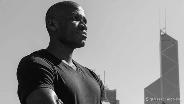 A black-and-white photograph of a muscular man with arms crossed standing in profile, with a watermark reading '© Masao Fast News' and a city skyline featuring a tall building in the background.