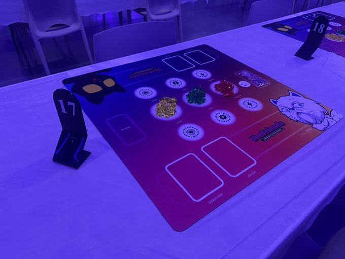A tabletop game mat labeled 'W-Origin' features colorful chips and cards, illuminated by purple lighting on a white table with black standees marked '17' and '18'.