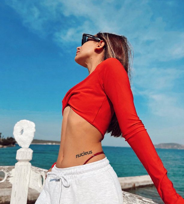 A woman in a red crop top and white pants with 'nucleus' tattoo visible on her hip, posing outdoors near the ocean.