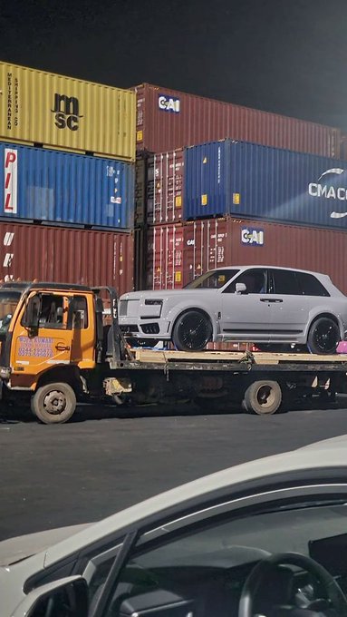 A white SUV being transported on a flatbed tow truck at a shipping port, with stacked shipping containers from MSC, CMA CGM, and PIL visible in the background.