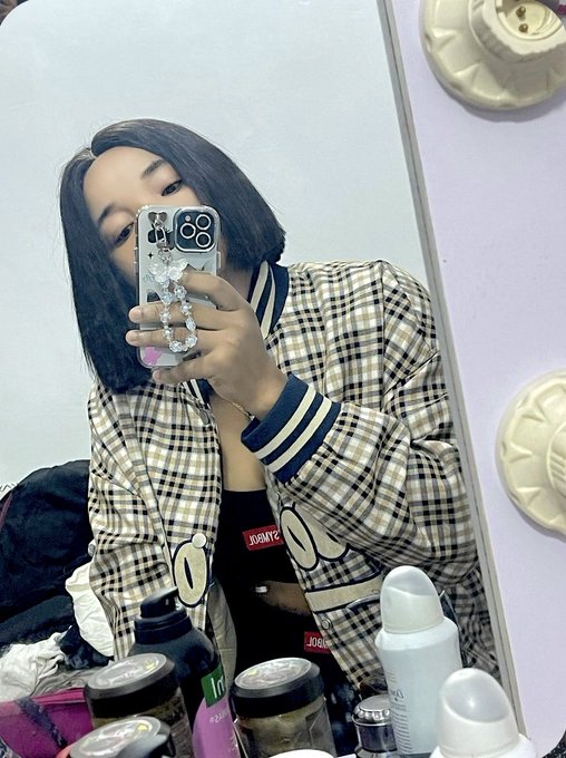 Mirror selfie of a young woman with short dark hair taking a photo with an iPhone decorated with charms, wearing a plaid jacket over a black top, with various bottles and items on a vanity in the foreground.