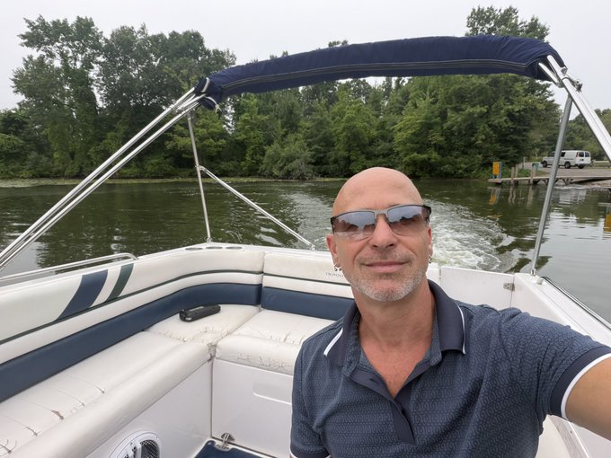 A bald man wearing sunglasses takes a selfie while driving a motorboat on a river lined with green trees.