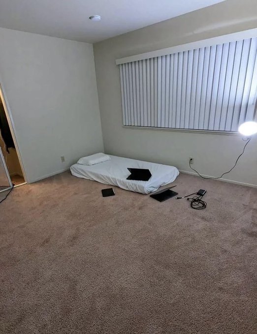 A sparse bedroom with a mattress on the floor, vertical blinds, and some electronics scattered on the carpet, humorously illustrating the meme that traders who gamble on memecoins often have lavish paper profits but live in minimalist, bare-bones conditions.