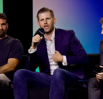 A photograph of Eric Trump, recognizable by his resemblance to Donald Trump, speaking into a microphone while wearing a navy blazer and white shirt at what appears to be a panel discussion or conference.