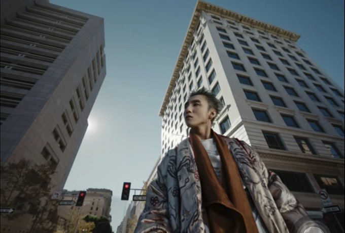 The image features a man wearing an oriental-style coat, standing in front of tall buildings with windows. The man appears to be looking up at something or someone above him, possibly admiring the cityscape or observing his surroundings. The photo is taken from a low angle, which emphasizes the height and grandeur of the buildings as well as the man's presence within this urban environment.