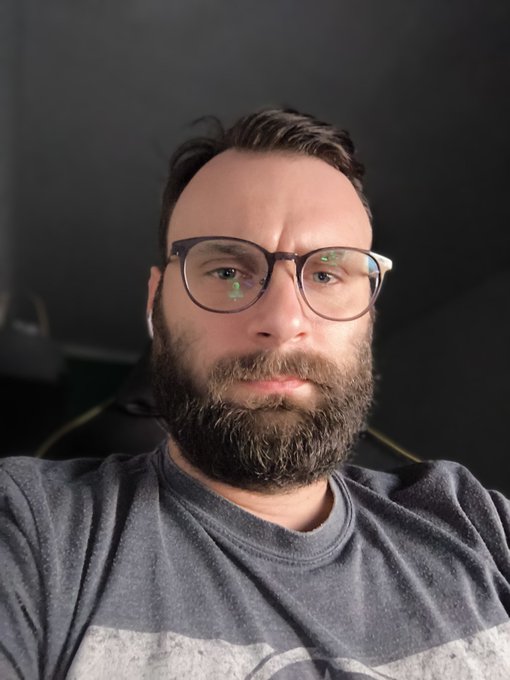 The image shows a man wearing glasses, a gray t-shirt with a graphic design, and a beard. The man appears to be looking directly at the camera while wearing his glasses. There are no texts or other objects visible in the image.