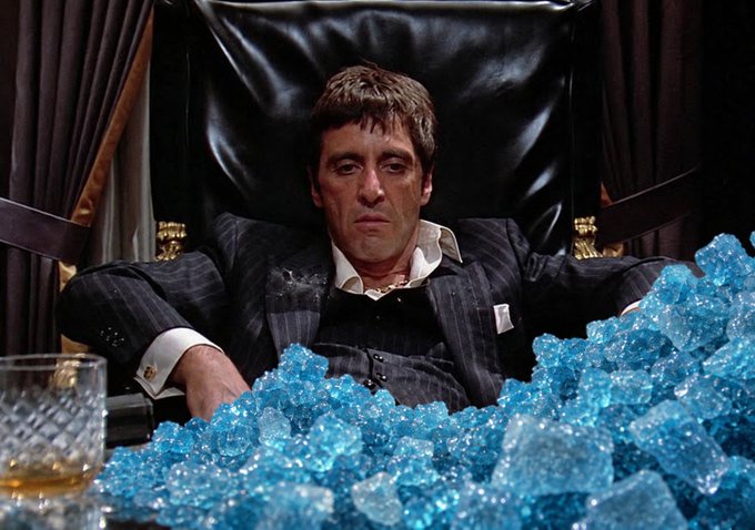 Al Pacino as Tony Montana sits in a black leather chair surrounded by a massive pile of blue crystals, wearing a dark pinstripe suit with a gold watch.