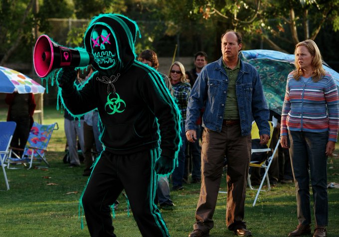 A composite image showing a glowing green-outlined hooded figure with a megaphone in the foreground, with people reacting in the background at what appears to be a park gathering.