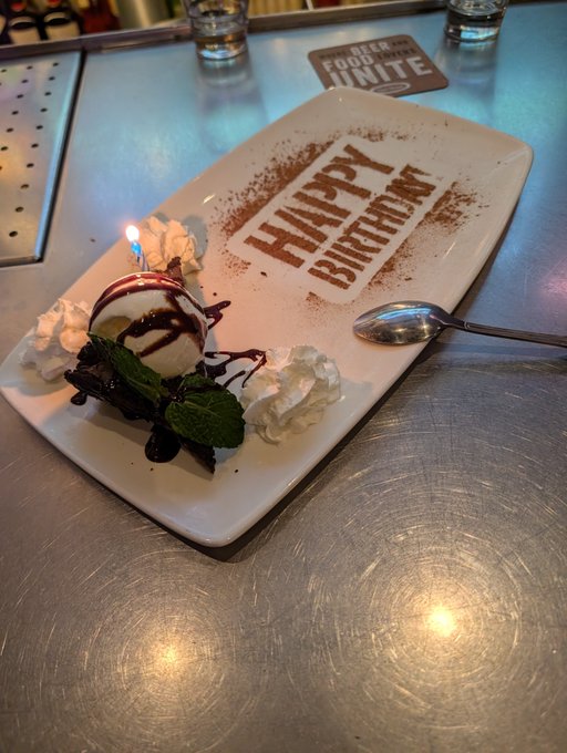 A rectangular white plate on a metal surface features an ice cream sundae with whipped cream and a lit candle, with 'HAPPY BIRTHDAY' written in cocoa powder on the plate.