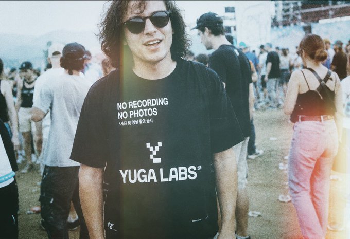 A man with curly hair and sunglasses wearing a black 'YUGA LABS' 'NO RECORDING NO PHOTOS' t-shirt at an outdoor festival.