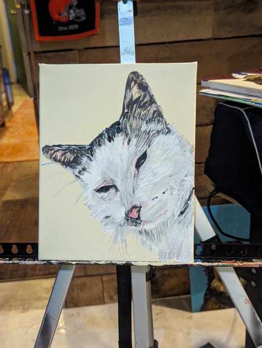 A textured painting of a white cat with dark ears and markings sits on a wooden easel in a studio setting.