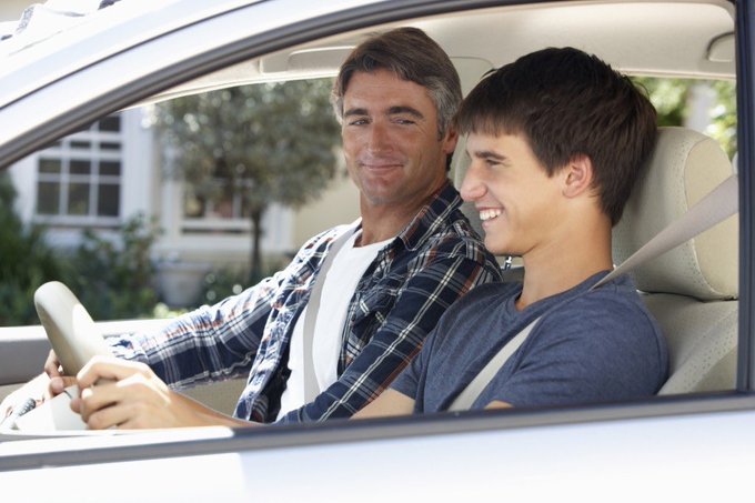 A smiling older man and a young teenage boy wearing seatbelts sit together in the driver's and passenger seats of a car.